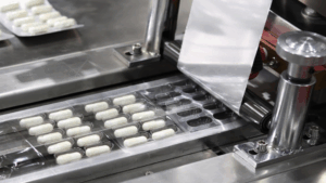 Máquina de embalagem tipo blister em linha de produção farmacêutica selando cápsulas brancas, destacando a importância da análise de extraíveis e lixiviáveis dos materiais para segurança do medicamento.