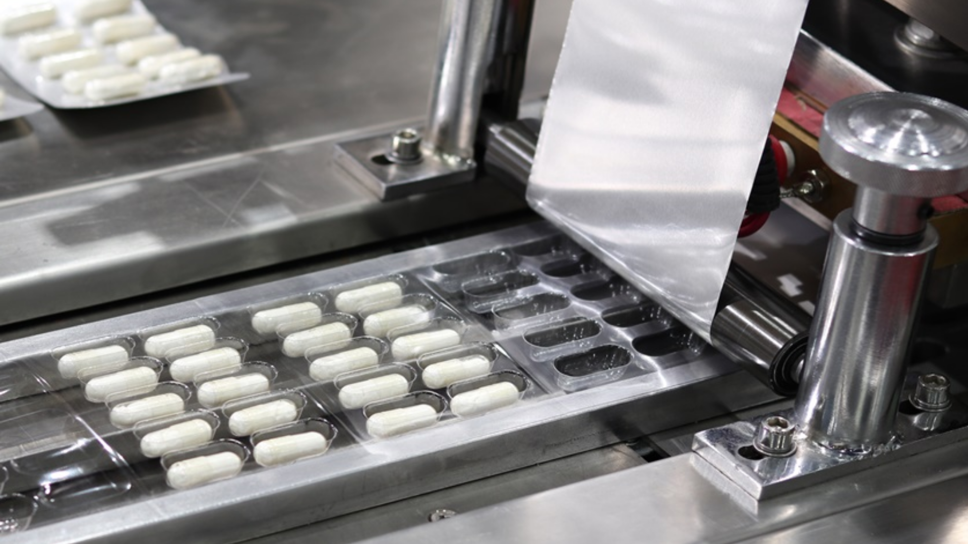 Máquina de embalagem tipo blister em linha de produção farmacêutica selando cápsulas brancas, destacando a importância da análise de extraíveis e lixiviáveis dos materiais para segurança do medicamento.