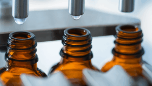 Frascos de vidro âmbar sendo envasados em linha de produção farmacêutica, ilustrando a etapa crítica onde a interação entre produto e embalagem pode gerar lixiviáveis.